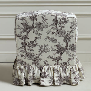 A ruffled fabric chair slipcover with a classic toile floral and bird pattern in dark gray on an off-white background, shown on a chair against a white paneled wall.