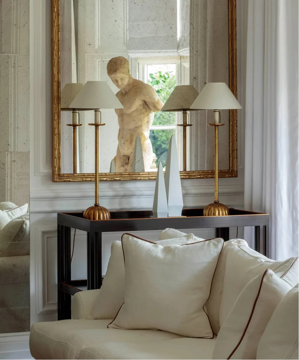 A large ornate gold-framed mirror hangs on a white paneled wall above a dark console table. The mirror reflects a classical-style stone bust sculpture, flanked by two brass table lamps with white drum shades.