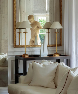 A large ornate gold-framed mirror hangs on a white paneled wall above a dark console table. The mirror reflects a classical-style stone bust sculpture, flanked by two brass table lamps with white drum shades.
