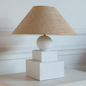 Table lamp with a beige woven shade on a white base against a light-colored wall.